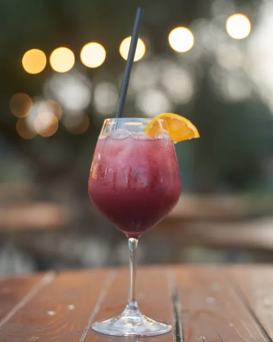 Un drink rosso con una fetta d'arancia su un tavolo di legno della Pizzina, con le luci accese sullo sfondo
