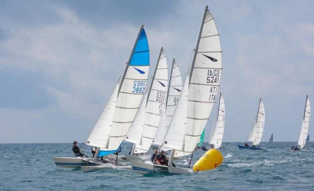 Catamarani in regata vicino a una boa gialla, con le vele bianche e una blu